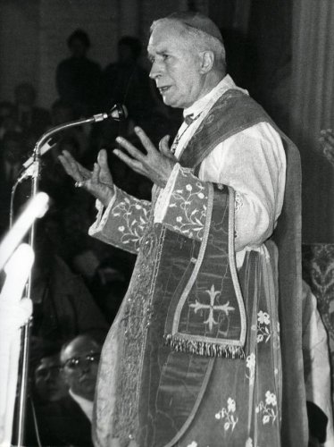 Vescovo anziano in abito liturgico parla al microfono, con mani aperte, durante una cerimonia religiosa.