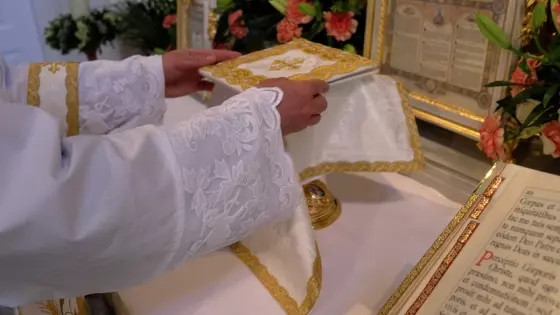 Persona in abito bianco ricamato sistema una tovaglia dorata su un altare decorato con fiori.
