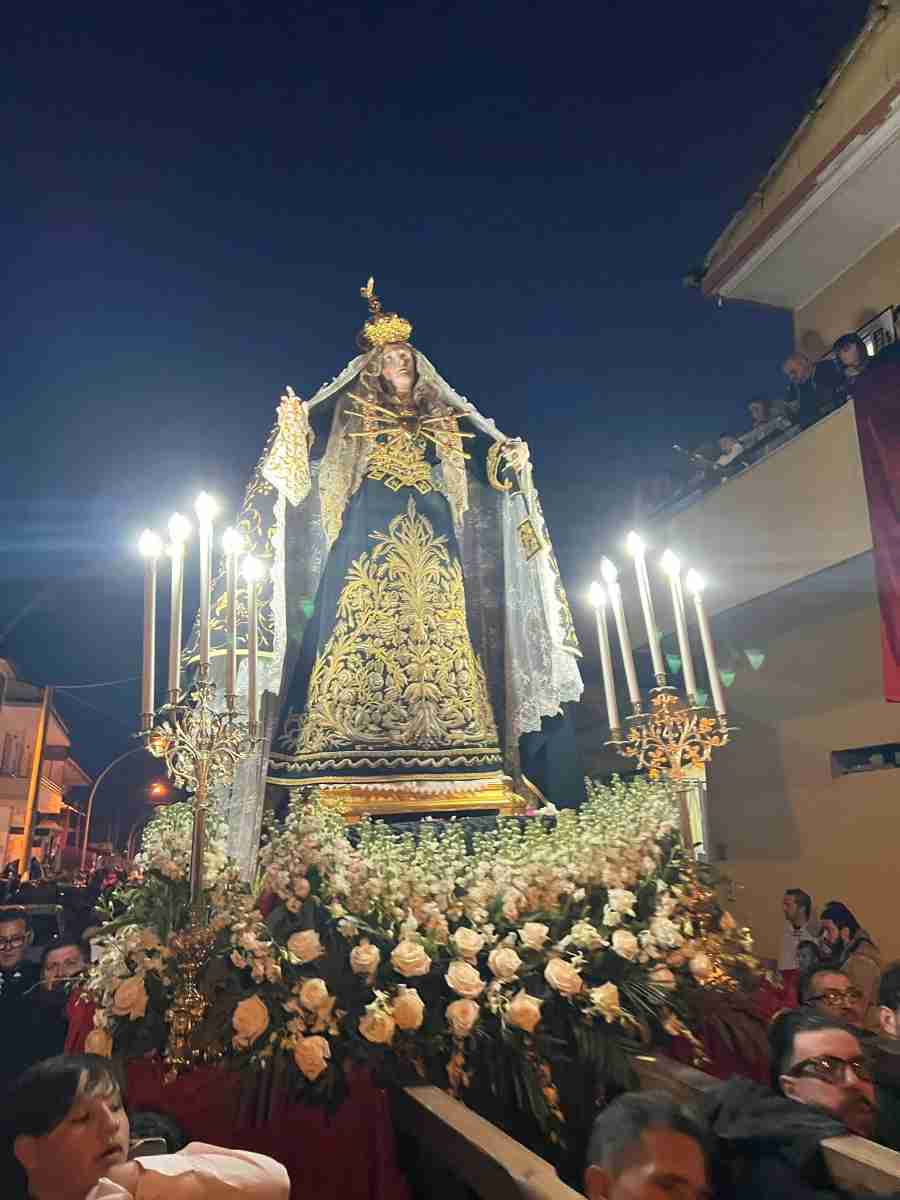 Statua religiosa dorata e vestita, illuminata da candele, circondata da fiori e folla durante una processione serale.